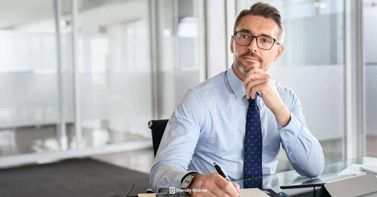 A professional middle-aged businessman with glasses sits at his office desk, looking thoughtful as he holds a pen to his chin.