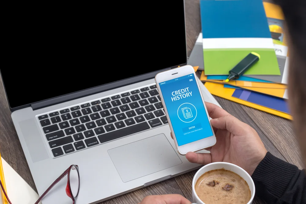 Person checking credit history on smartphone next to laptop and coffee