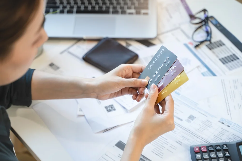Person holding multiple credit cards over a messy desk filled with overdue bills and a calculator