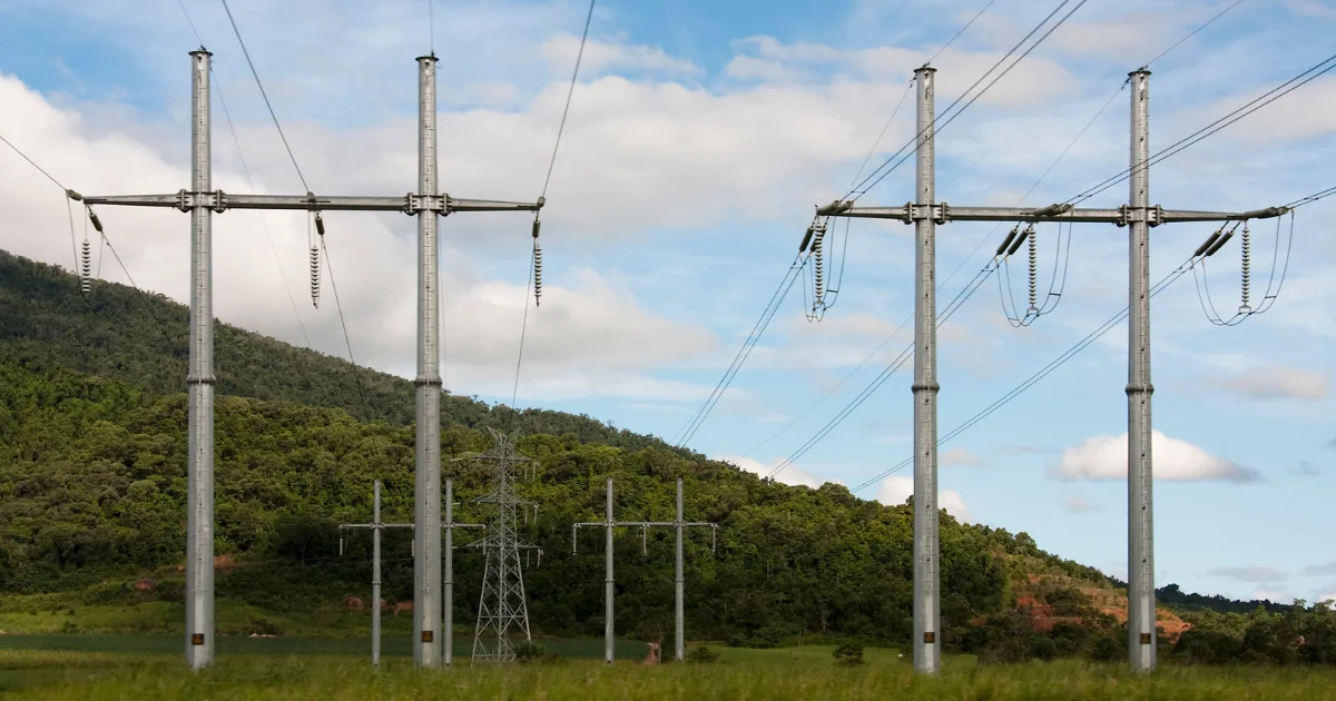 Tall metal electricity transmission towers holding high-voltage power lines running across a lush green hill and field under a blue sky with scattered clouds.