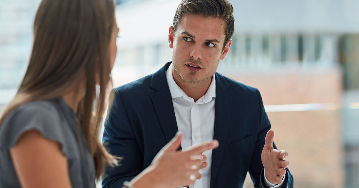 A male financial consultant in a dark blazer and white dress shirt gesturing with his hands while speaking seriously to a female client during a meeting.