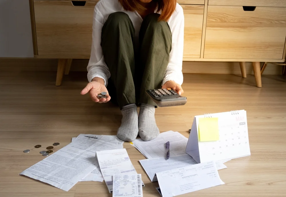 Woman sitting on floor surrounded by bills, calendar, coins, and a calculator, appearing financially stressed