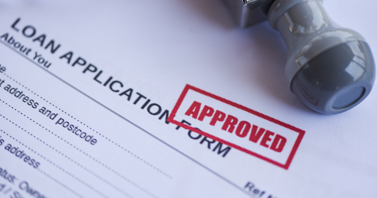 A close-up of a paper loan application form featuring a bold red ink stamp that reads "APPROVED" next to the handle of a desktop stamp tool.