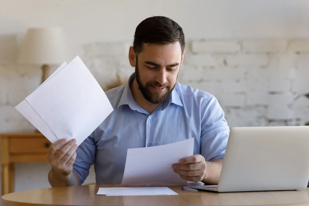 Smiling man reviewing paperwork at a desk with a laptop in a bright home office setting