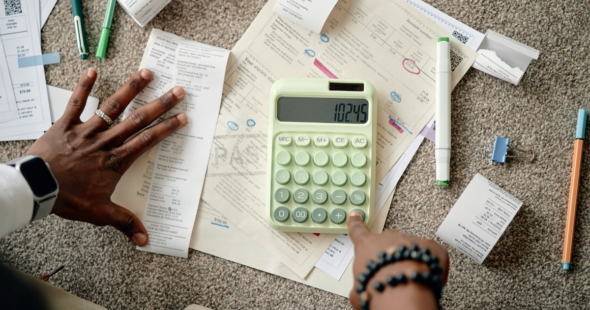 A top-down view of a person using a light green calculator displaying "102.45" to tally up pile of utility bills, receipts, and invoices scattered on a carpet.