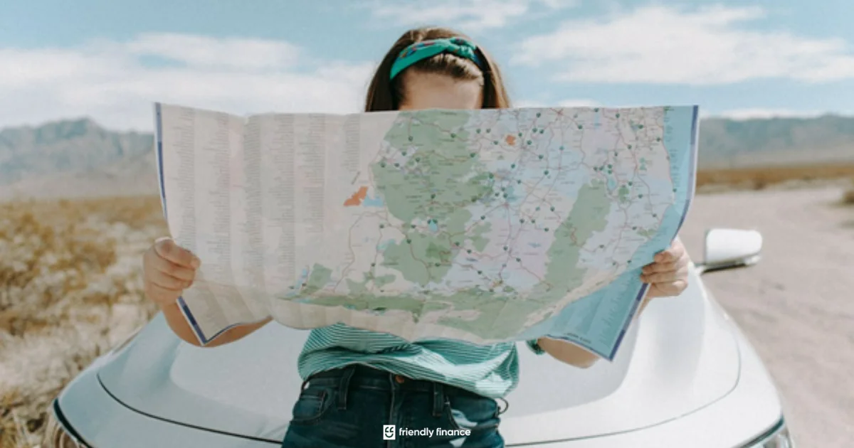 A person on a road trip leans against the front of a car while reading a large paper map, with a sunny landscape in the background.