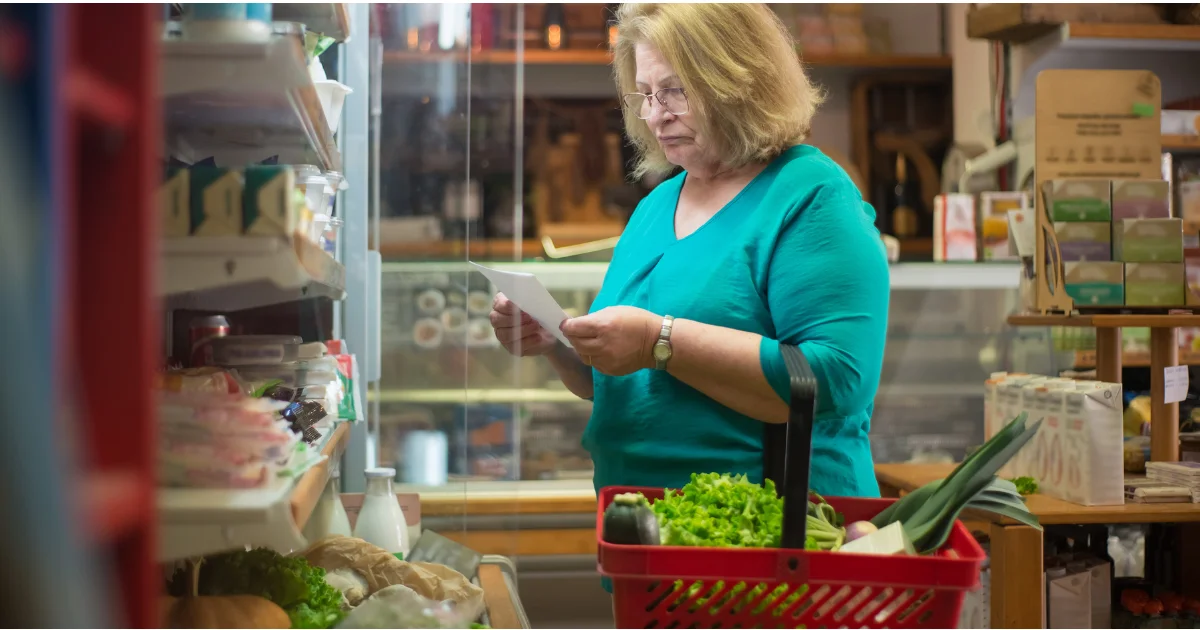 An elderly woman with glasses standing in a supermarket aisle, holding a red shopping basket filled with fresh leafy greens while reviewing a grocery shopping list.