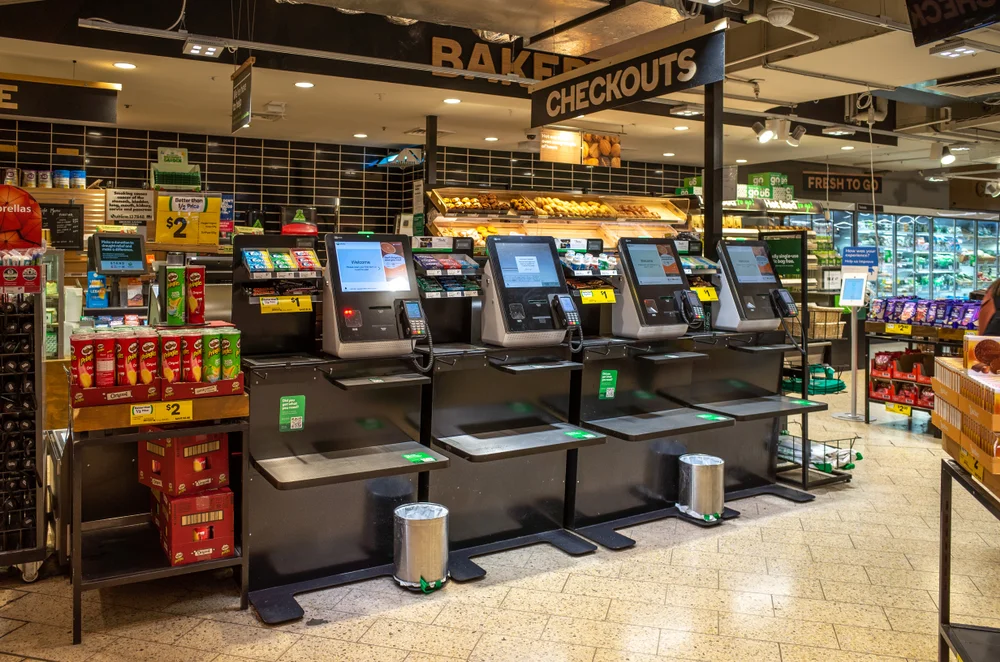 Self-checkout stations inside a supermarket, with bakery goods and grocery items in the background