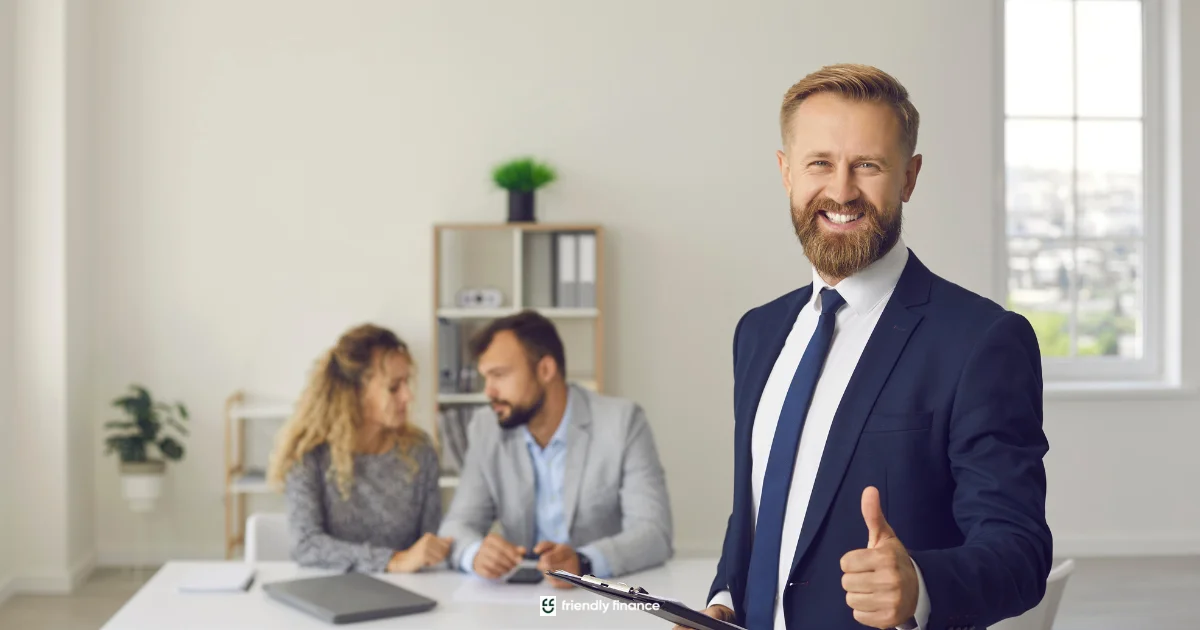 Smiling loan broker giving thumbs up while clients review loan documents in the background.