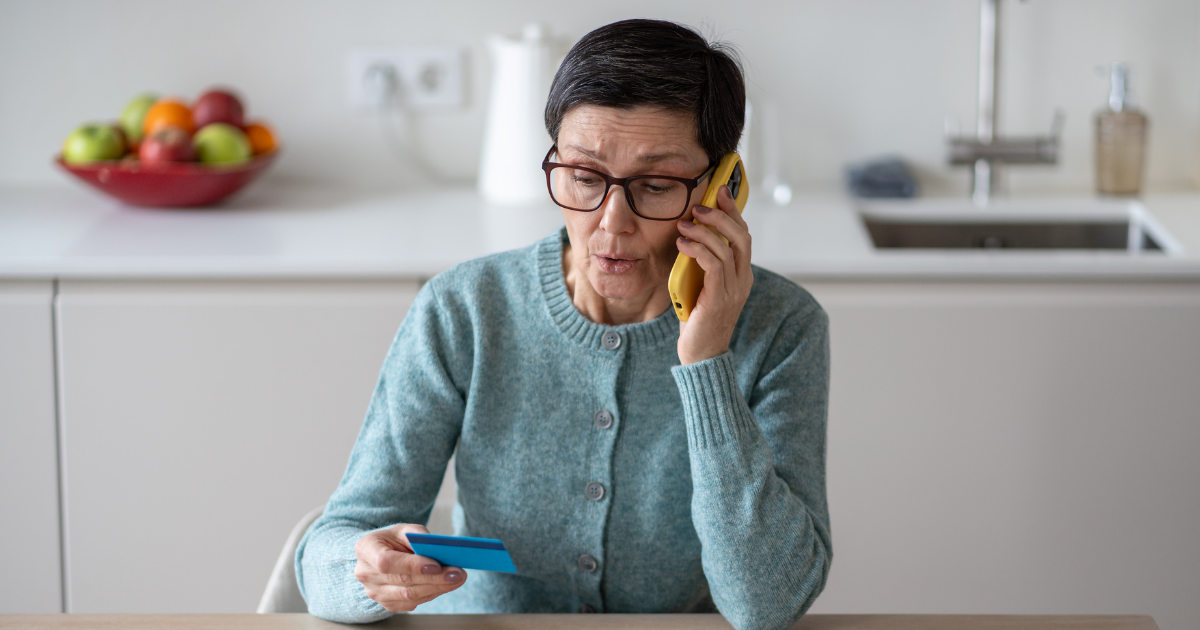 An older woman with short hair and glasses sitting at a kitchen table, holding a blue credit card while talking on a yellow mobile phone with a concerned expression.